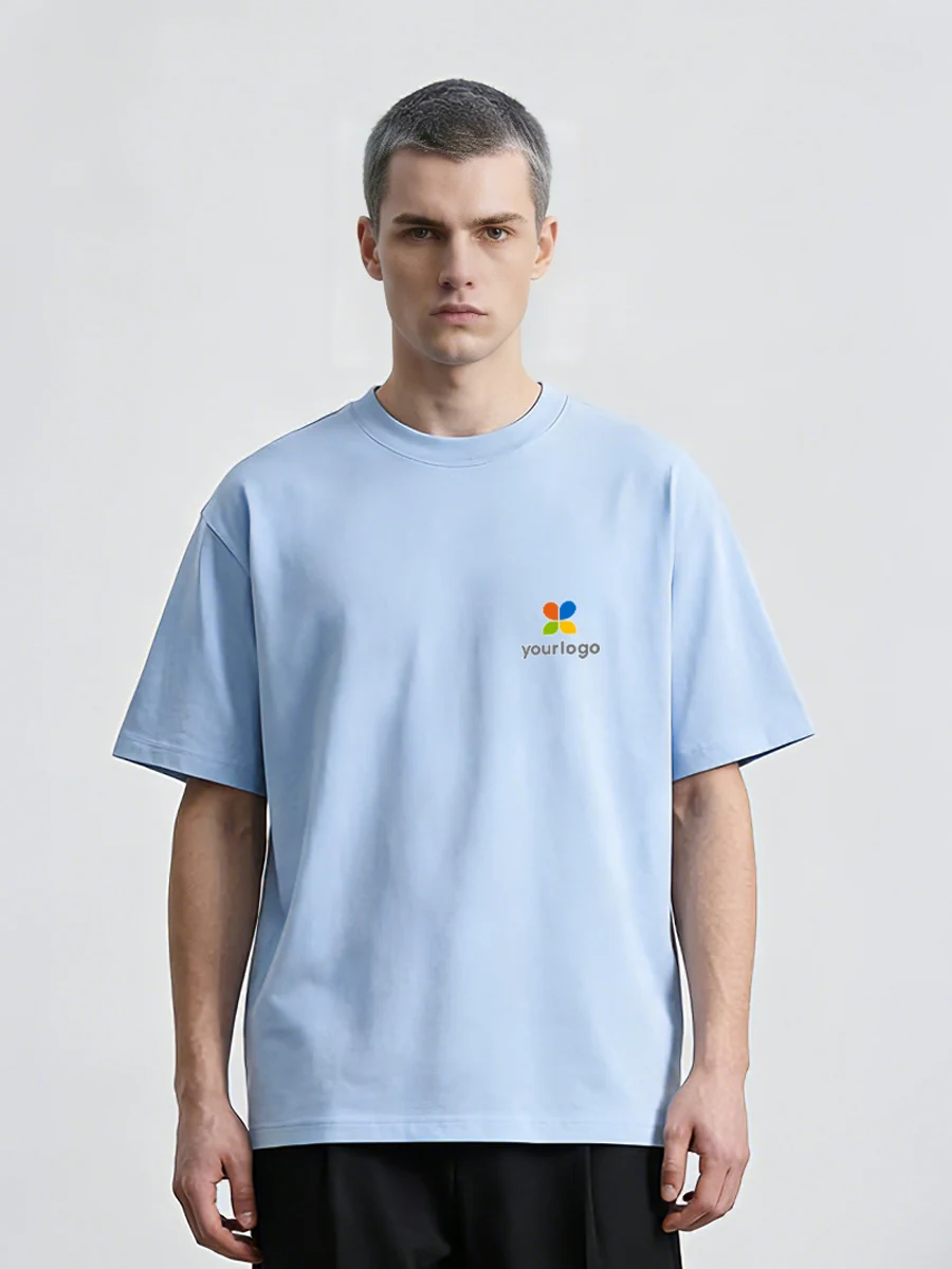 Man in a sky blue shirt with a rainbow logo stands against a white wall. The minimalist, figurative design features tech wear aesthetics and a bourgeois vibe. The unisex short-sleeve t-shirt is made of heavyweight cotton blend, moisture-wicking and quick-drying, ideal for campus wear and daily commutes.